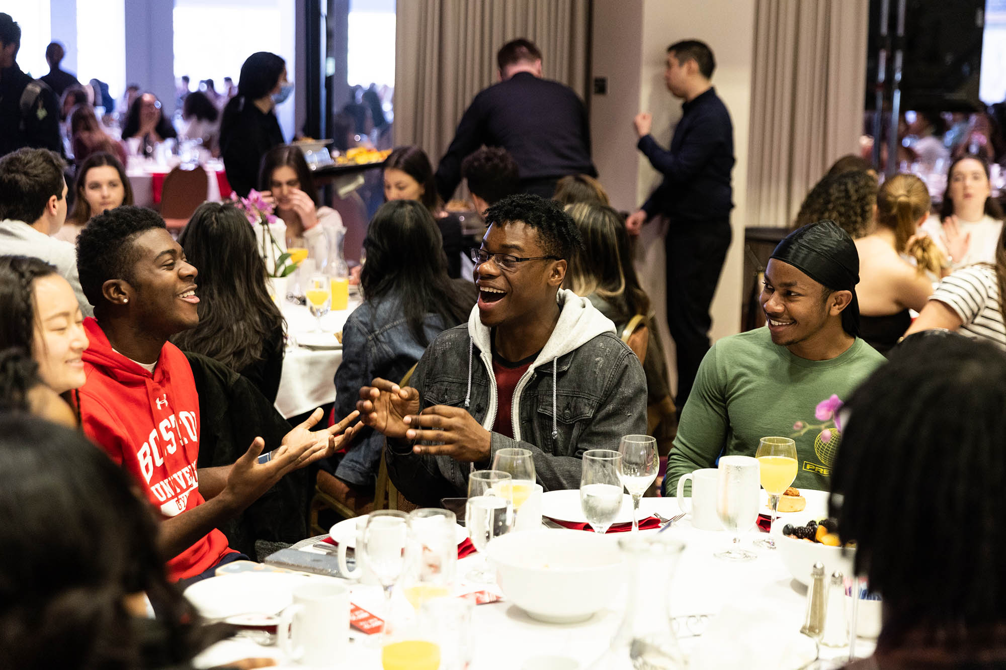 Photo: Three young Black men dressed in casual clothes laugh and speak in excitement to each other as they sit at a large round table filled with plates. Other students sit around them at similar tables.