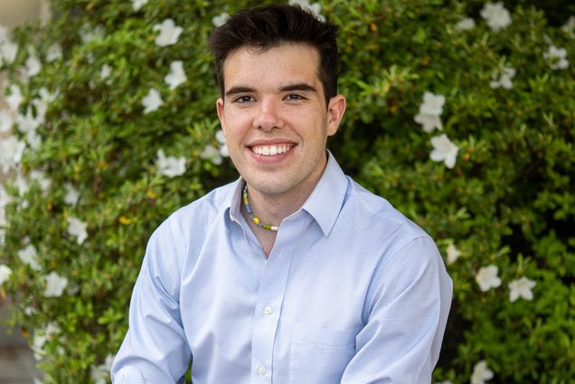 Photo: Sumner Jones, a young white man with dark brown hair and wearing a light blue, long-sleeved collared shirt, smiles and poses in front of a large, flowering green bush.