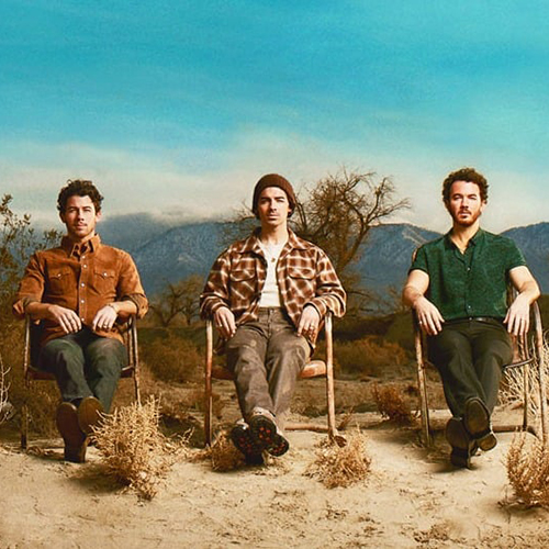 Photo: The Jonas Brothers sit in three chairs side by side in a desert during a cloudless, sunny day.