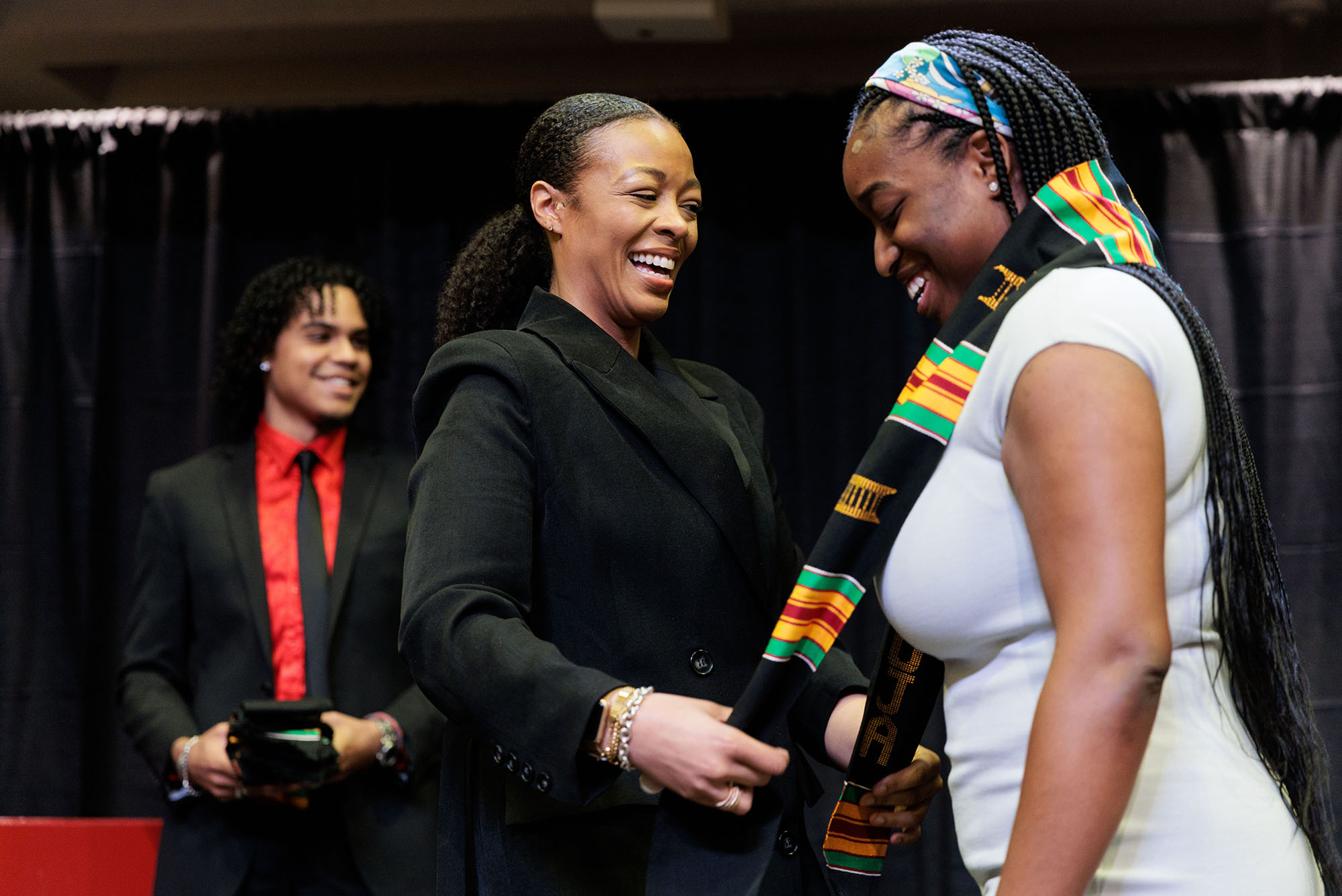 Photo: (standing from left) Joshua Diamond (CAS'22), and Professor Saida Grundy, Director of Admissions, congratulate Jada Warmington (QST'23) during Rite of Passage. A Black woman (center) wearing an all black suit ensemble smiles as she places a black, yellow, red, and green graduation stole around a young Black woman's shoulders (right).