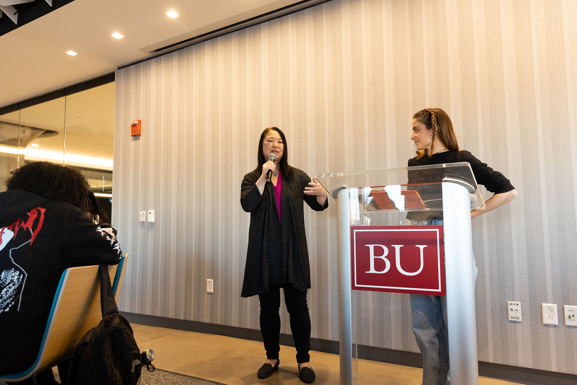 Photo: Two individuals--Director of the Newbury Center, Maria Erb, left, introduces Vicky Plasencia, right-- are standing at a BU podium as Erb introduces Plasencia to the crowd. Erb wears a black outfit with a long balzer and Plasencia wears an all black outfit.