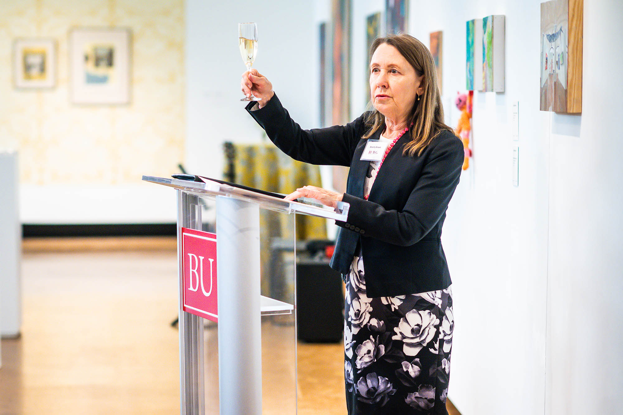 Photo: Dr. Beverly Brown, a white woman wearing a white blouse, black blazer, and black and white floral skirt, holds up a glass of champagne as she stands at a glass podium and addresses a crowd during a reception.