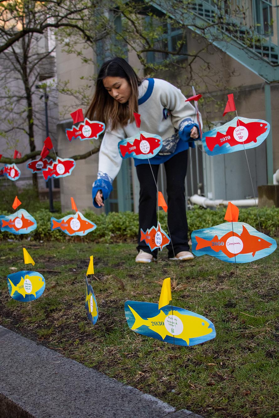 Photo: A young woman plants stakes with paper fishes in a grassy area for a World Ocean Day Installation. The fishes are colorful, a mix of red, orange, and yellow and the young student wears black yoga pants and a gray sweatshirt.