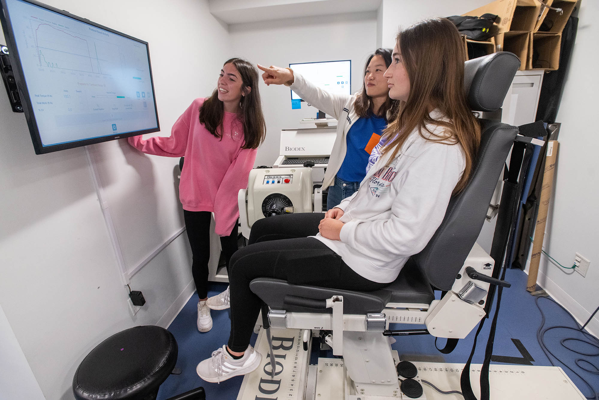 Photo: Three woman look at a screen depicting data. One, in the chair wears black leggings and a white sweatshirt. The next, an Asian woman, wears a blue shirt and a white button up. The last and closet to the screen, a white woman, wears a pink shirt and leggings.