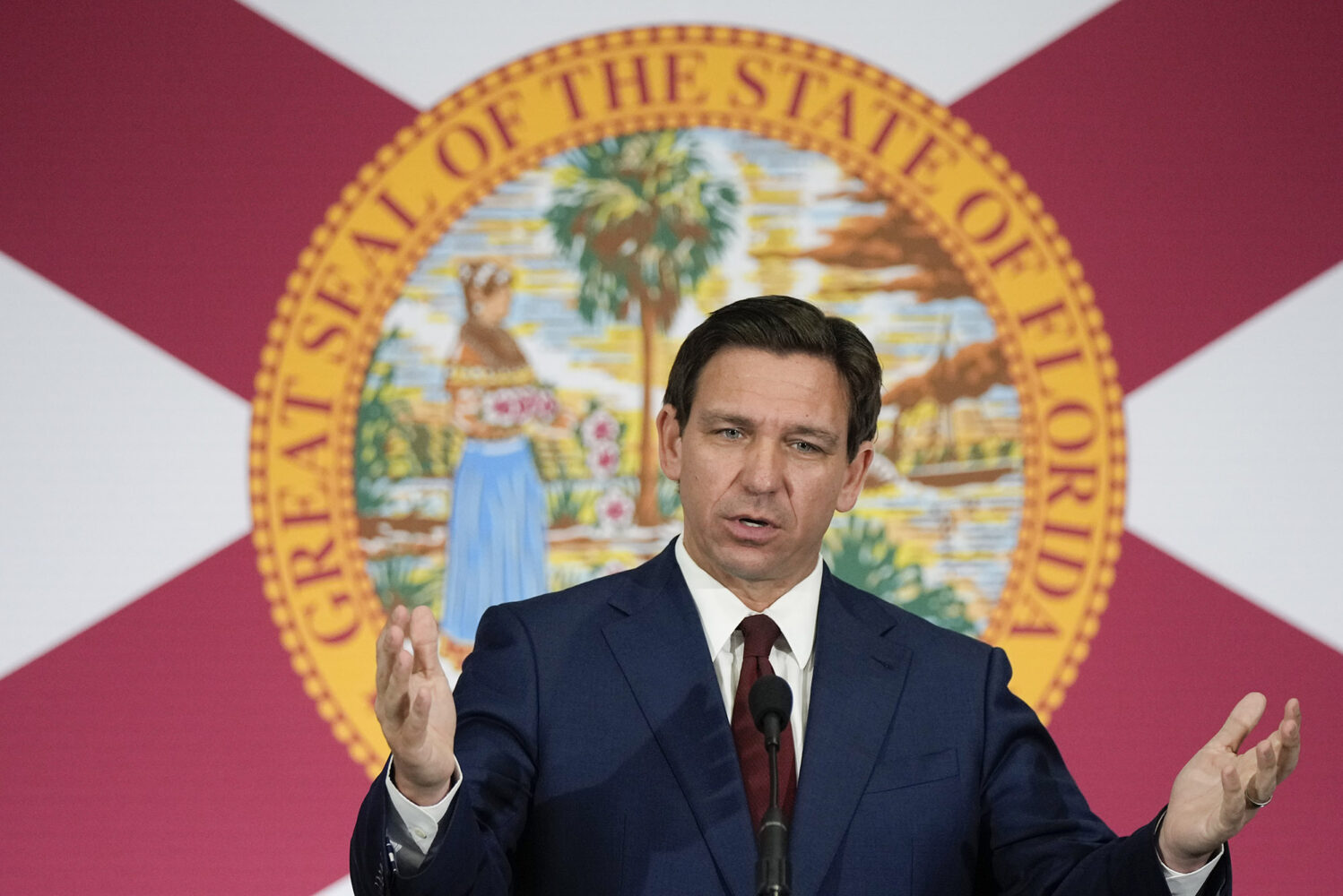 Photo: Ron DeSantis, a white man, stands in front of a banner with Florida's emblem addressing a crowd. He wears a basic navy suit with a red tie.