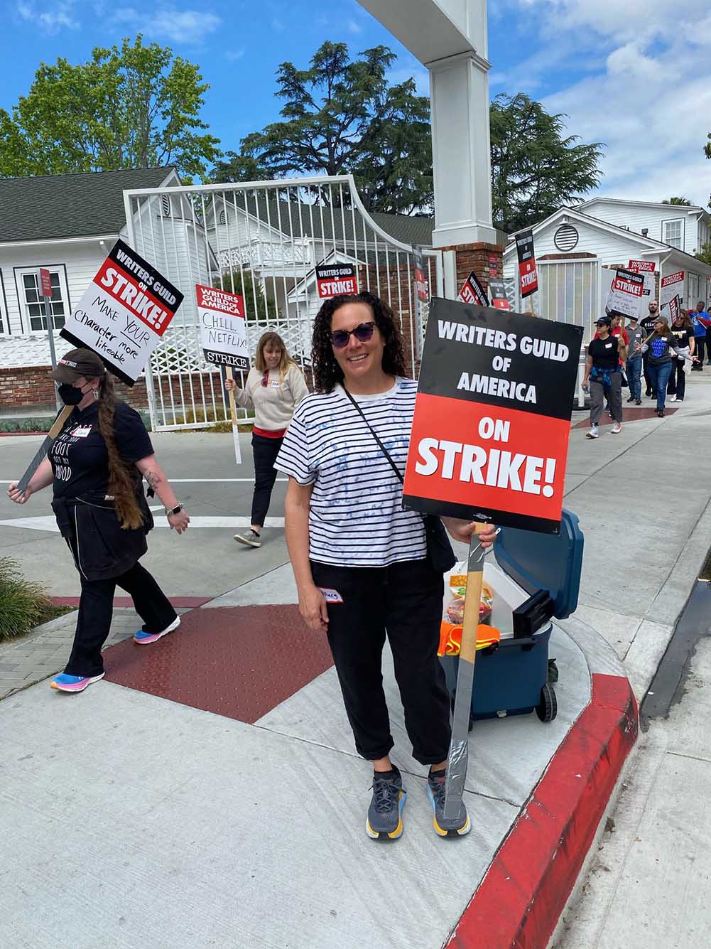 Photo: Hilary Weisman Graham (COM’92), a white woman wearing sunglasses, a blue-striped white shirt and black pants, smiles and poses with a picket sign that reads "Writers Guild of America ON STRIKE!"