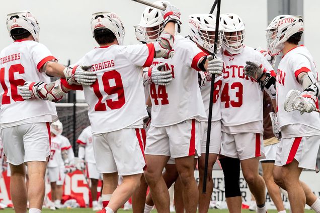 Photo: A group of BU lacrosse team members celebrate in a huddle. They are wearing their white uniforms with red detailing and are playing on in inside field.