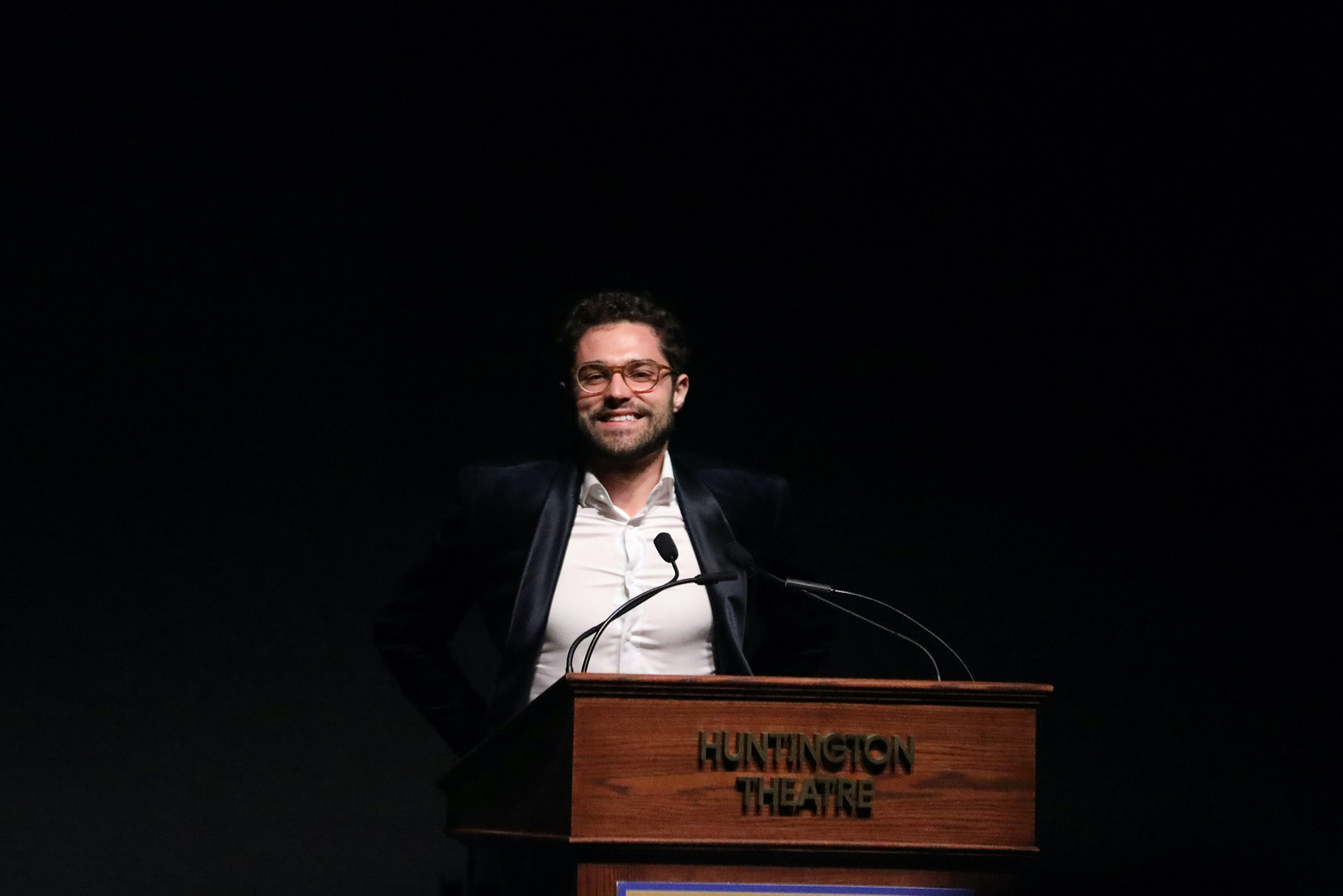Photo: Mishka Yarovoy (CFA’23) accepts the Elliot Norton Award. A white man wearing glasses, a white collared shirt, and velvet black blazer smiles as he stands behind a wooden podium that reads "Huntington Theater".
