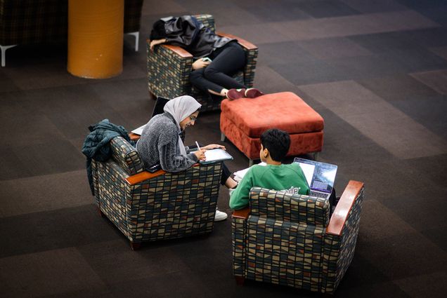 Photo: Three students sit in chairs around an ottoman. The two chairs in foreground, the students are discussing something about their study materials. The chair in the background features a student curled up with a coat over them, completely passed out. The first student in the foreground wears a green t-shirt, the second student wears a gray hijab and a shirt. The chairs are orange and a dark pattern.