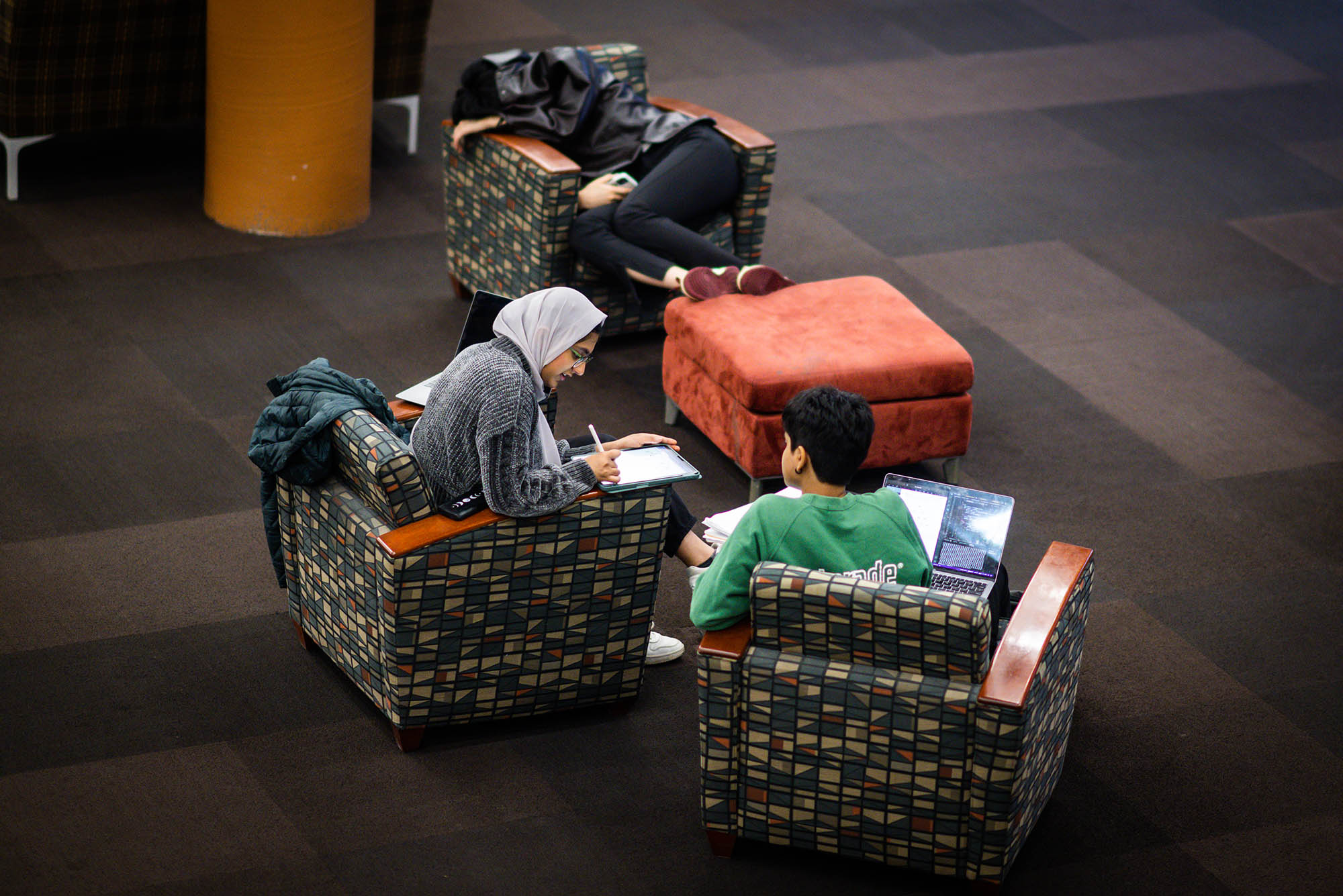Photo: Three students sit in chairs around an ottoman. The two chairs in foreground, the students are discussing something about their study materials. The chair in the background features a student curled up with a coat over them, completely passed out. The first student in the foreground wears a green t-shirt, the second student wears a gray hijab and a shirt. The chairs are orange and a dark pattern.
