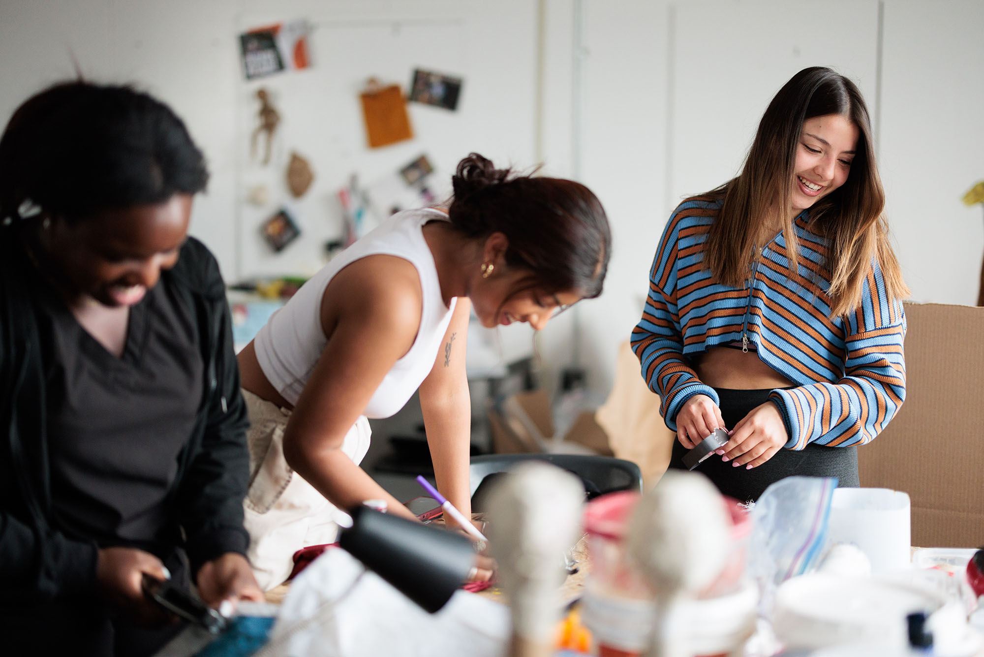 Photo: Three women create and put final touches on puppets. From left, a Black woman in all black. In the middle, an hispanic woman in all white and cream. On the left, a white woman with a striped shirt.