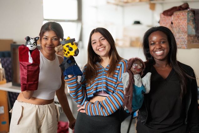 Photo: Three young women hold up puppets on their right hand and smile into the camera. From left, she has short brown hair and wears a monochromatic tan outfit. In the middle, a white woman with long brown hair and a stripped shirt. On the right, a Black woman with shoulder length straight hair. She wears an all black outfit.