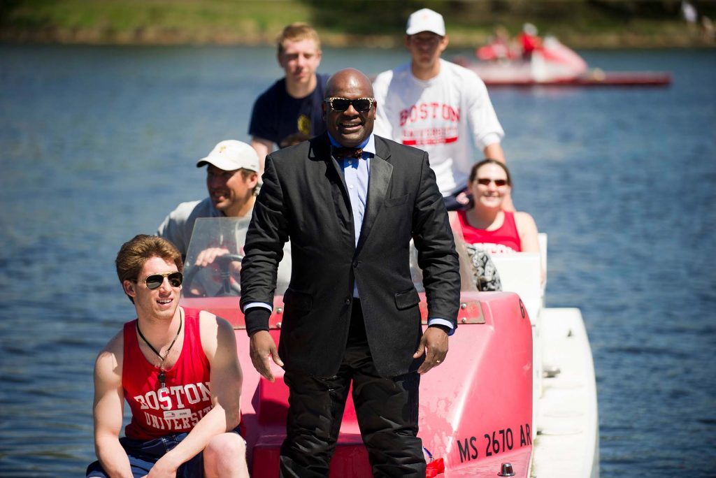 Photo of Dean Elmore, in a black suit, standing at the bow of a launch boat, smiling. Students in red jerseys stand on the boat behind him. They are on the Charles and Elmore is preparing to jump in.