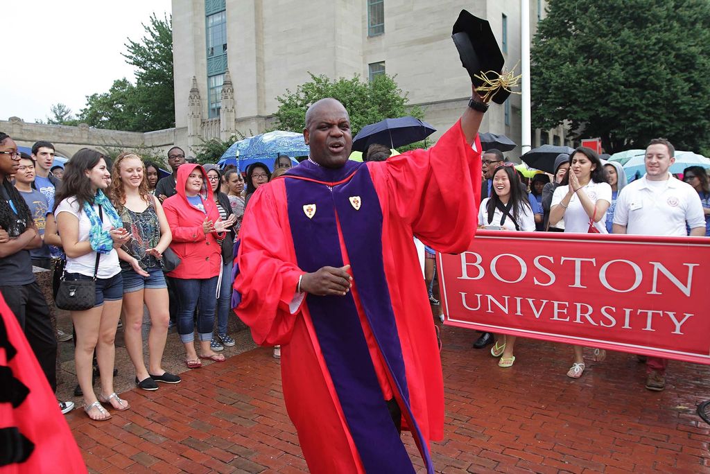 Photo of Ken Elmore at the front of the matriculation procession along Comm Ave. He wears a red robe, purple sash and raises a black hat in the air. Students holding a red BU sign can be seen at right.