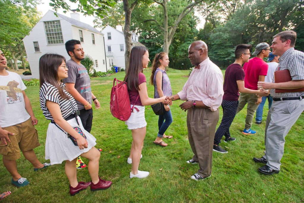 Photo of Ken Elmore, a middle aged Black man with a shaved head, center right, in a pink shirt and brown pants, shaking hands with incoming freshmen outside on a lawn. About six or seven people stand around him.