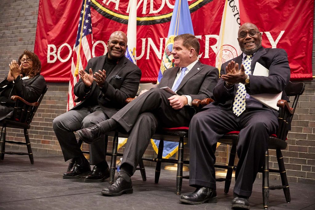 Elmore sits on stage with former Boston Mayor Marty Walsh, a white man in a grayish suit, and an older Black gentlemen in a suit who is seen clapping. Elmore smiles and wears a black suit and claps his hand. A Boston University flag is seen behind them as they sit on stage during an MLK celebration event at Metcalf.