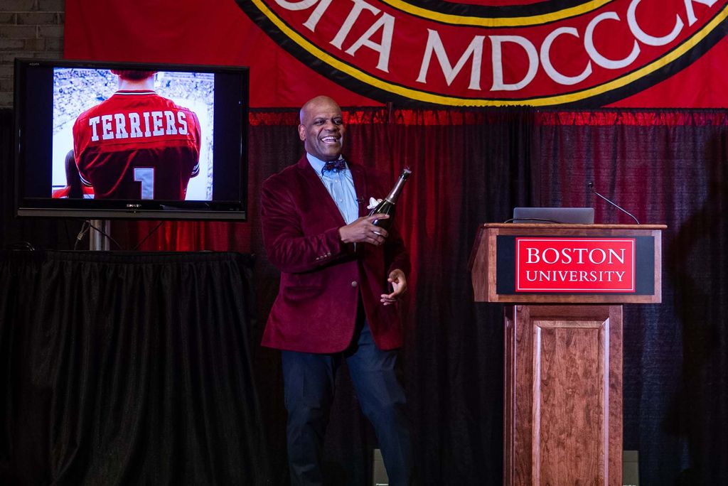 Photo of Ken Elmore, in a red velvet blazer, holding a champagne bottle in his left hand and smiling as he presents on stage during senior breakfast. At left, a TV with a photo of a hockey player is seen. At right, a podium with a red Boston University logo.