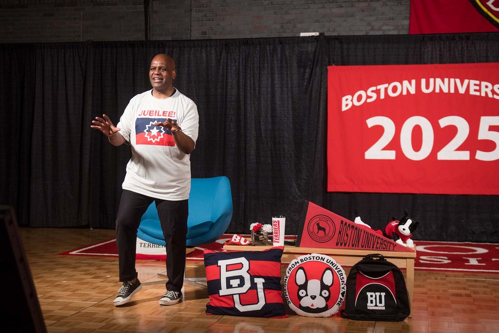 Dean Elmore, seen on stage at Metcalf, during a record of freshmen orientation. He wears a white t-shirt with a juneteenth flag and the word "Jubilee" written on it. Behind him hangs a banner with "Boston University Class of 2025" on it. On stage, to his left, is a organized pile of BU swag, including a flag , pillows and backpacks.
