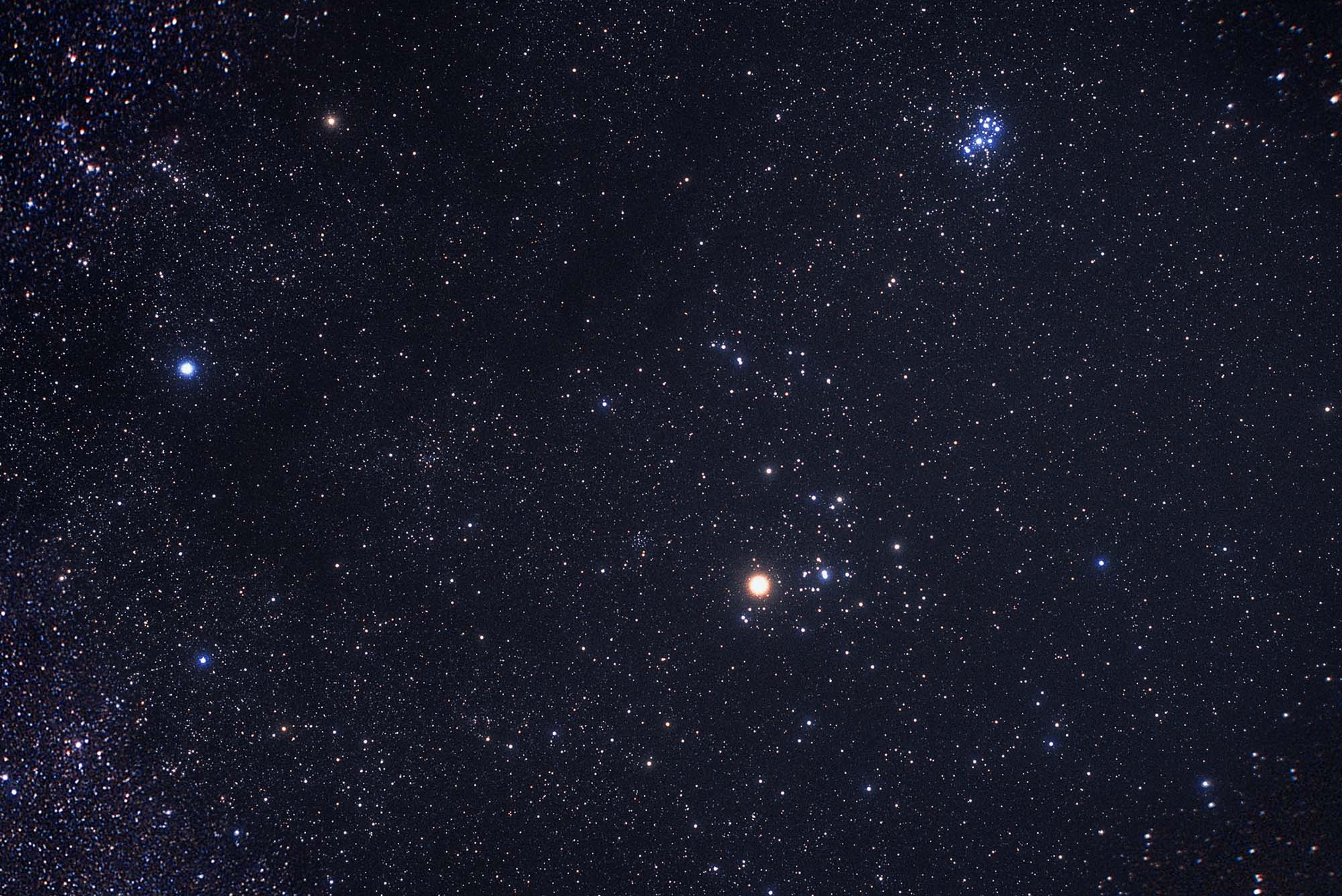 Photograph of the constellation Taurus made by the avid astrophotographer Akira Fujii. In the photo, tiny white dots, stars, fill the image, with a brighter, reddish dot towards the bottom right, which is Taurus.