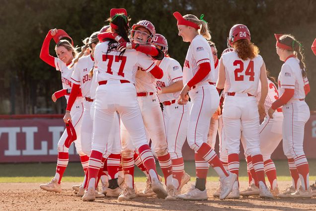 Photo: BU's softball team celebrates in a huge huddle, outside on the field. They were their white and red uniforms, the red be the detailing and numbers. Some embrace in a hug on the left portion of the screen, two with helmets and others with visors. It seems to be midday.