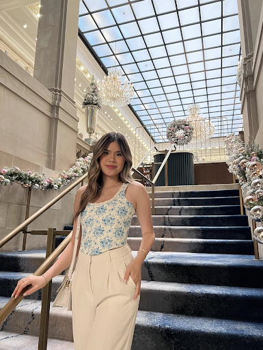 This is a photo of Boston University graduating senior Kylee Nguyen. She is in a blue-floral top with white pants on, standing on a stair case of a big building that has Christmas decor up. Photo credits: Kylee Nguyen (COM'23).