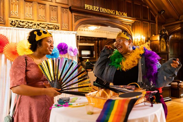 Photo: Two young Black women smile and laugh as they wear golden and rainbow accessories. The table and room around them are decorated with rainbow flags and accessories, as well.