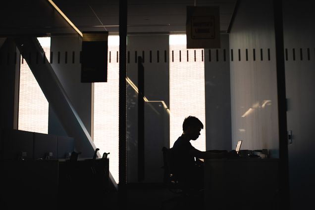 Photo: The silhouette of a student sitting and wearing glasses is shown. They sit in front of a large window with golden hour lighting as they study.