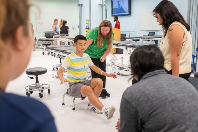 Photo: Henry Di, an Asian young man wearing a light blue, yellow, and white triped shirt and grey shorts, prepares himself to stand up from a short stool during a lab exercise. He sits with one leg held straight in front of him and arms out for balance. To his right, Clinical assistant professor Sorcha Martin, a white woman with long brown-blonde hair and wearing a green shirt and black pants, guides him with a hand on his back as she instructs other student onlookers.