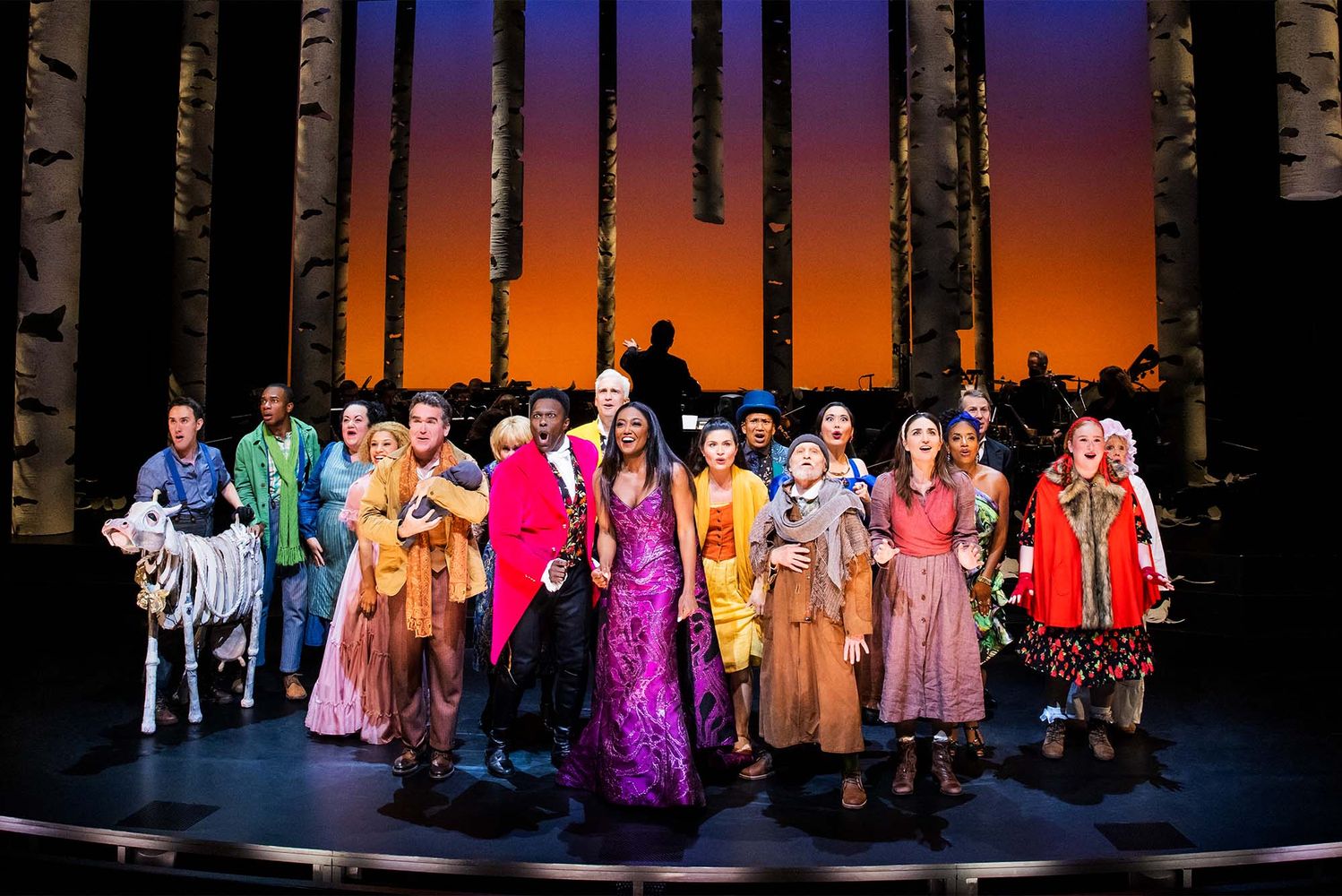 Photo: A large cast of people in costumes stand on a stage with a red background. They are wearing dresses and suits in various colors.