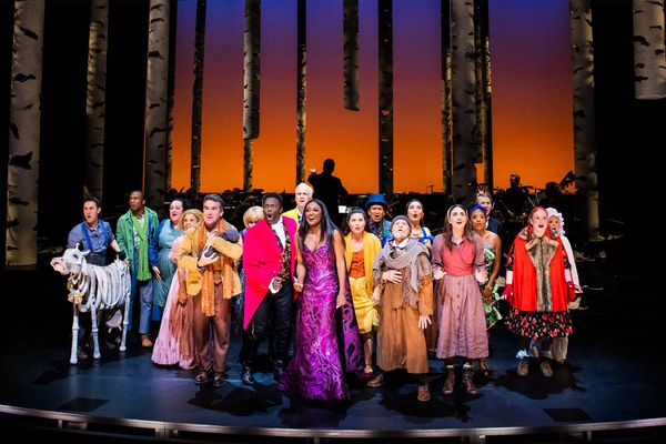 Photo: A large cast of people in costumes stand on a stage with a red background. They are wearing dresses and suits in various colors.