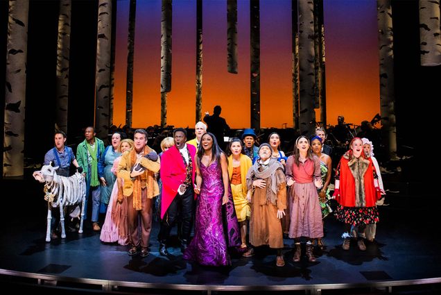 Photo: A large cast of people in costumes stand on a stage with a red background. They are wearing dresses and suits in various colors.