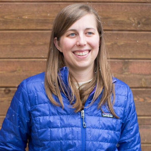 Photo: Alyssa Pierson, a white woman with long, light brown hair and wearing a blur puffer jacket, smiles and poses in front of a wooden plank wall.