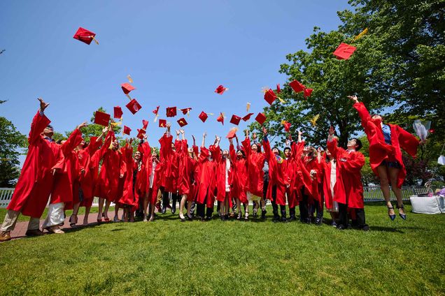 Photo: Lined up with their ceremonial cap and gown, some throwing their caps up in the air. The day is bright and sunny and the grass they stand on is green.