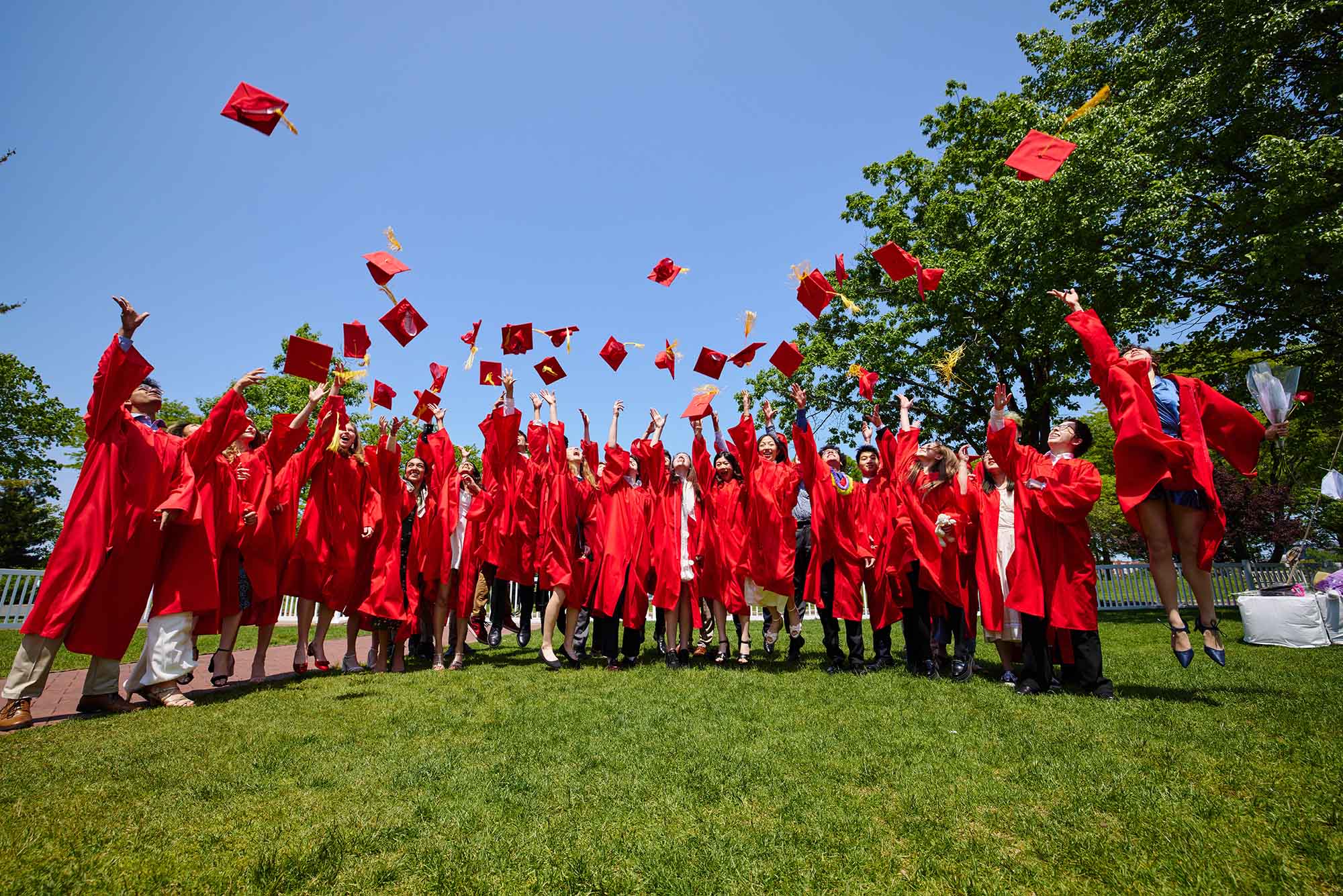 Photo: Lined up with their ceremonial cap and gown, some throwing their caps up in the air. The day is bright and sunny and the grass they stand on is green.