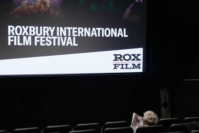 Photo: A older person with white hair sits alone in the front of a movie theater. The text on screen reads "Roxbury International Film Festival"