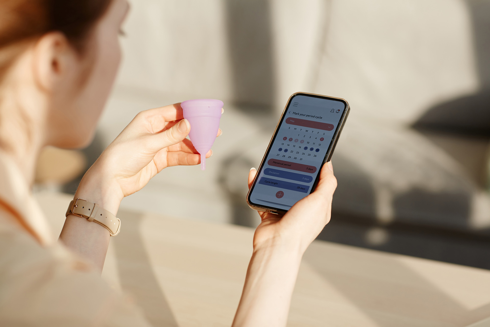 Close up photo of a young white woman calculating menstrual cycle using a generic mobile app and holding reusable menstrual cup cup on her other hand.