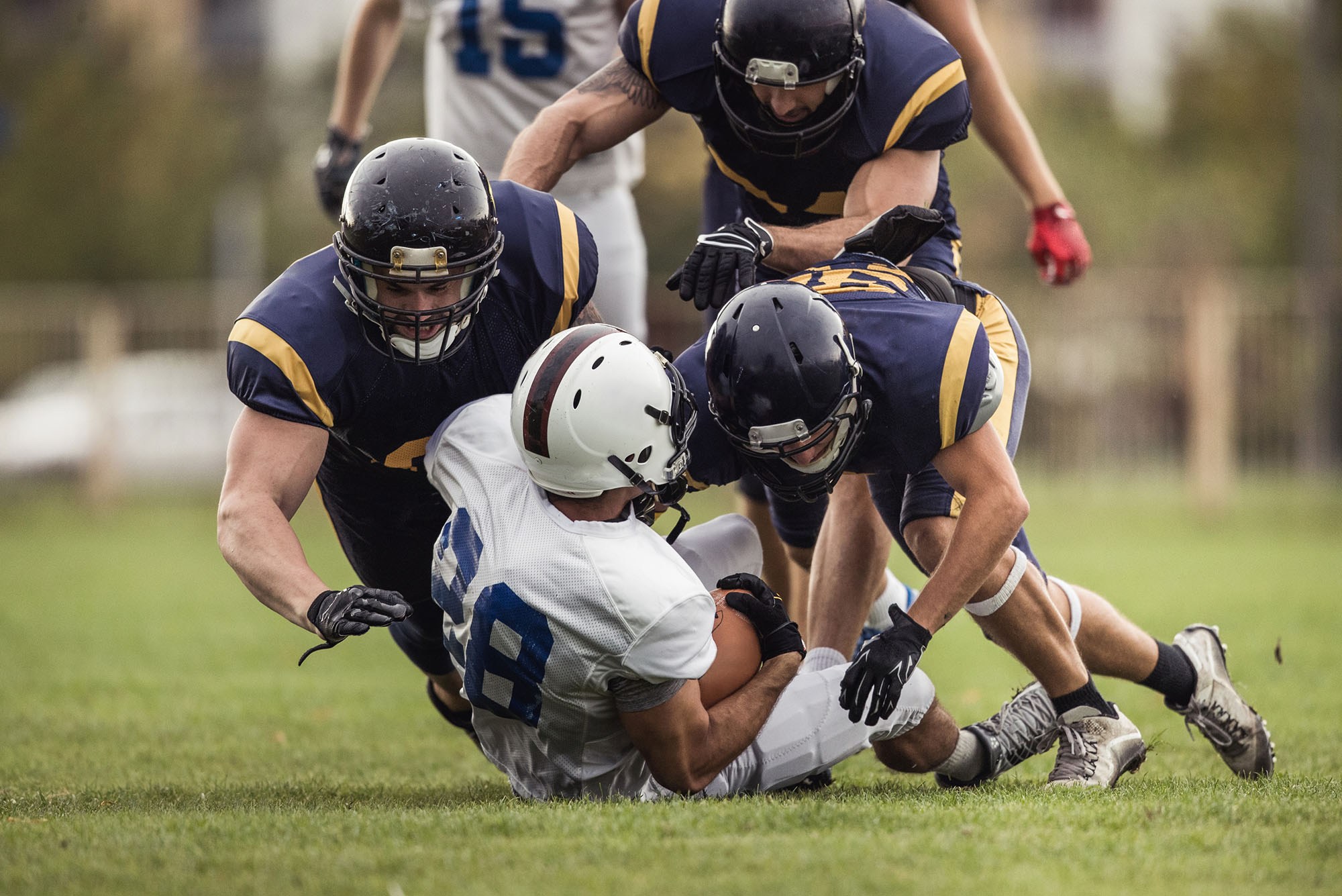 Photo: Group of American football players mid-game. Team member wearing black football uniforms and helmets are shown as they tackle a player holding the football and waring a white football uniform and helmet.