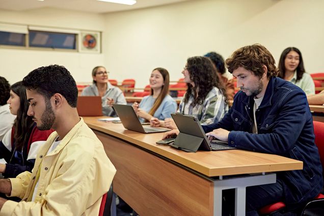 Photo: College students sit in a tiered college classroom as they work on laptops and chat with each other.