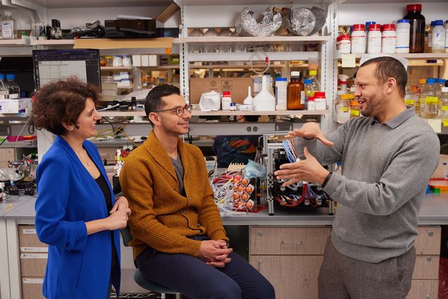 Photo: College of Engineering professors (from left) Rabia Yazicigil, a white woman with short curly hair and wearing a black blouse, oversized blue blazer, and black pants, Mo Khalil, A tan man wearing glasses, brown sweater, and black pants, and Doug Densmore, a light-skinned Black man wearing a long sleeve collared grey shirt and black pants in a lab. Yazicigil and Mo Khalil look to Densmore as he talks and gesticulates.