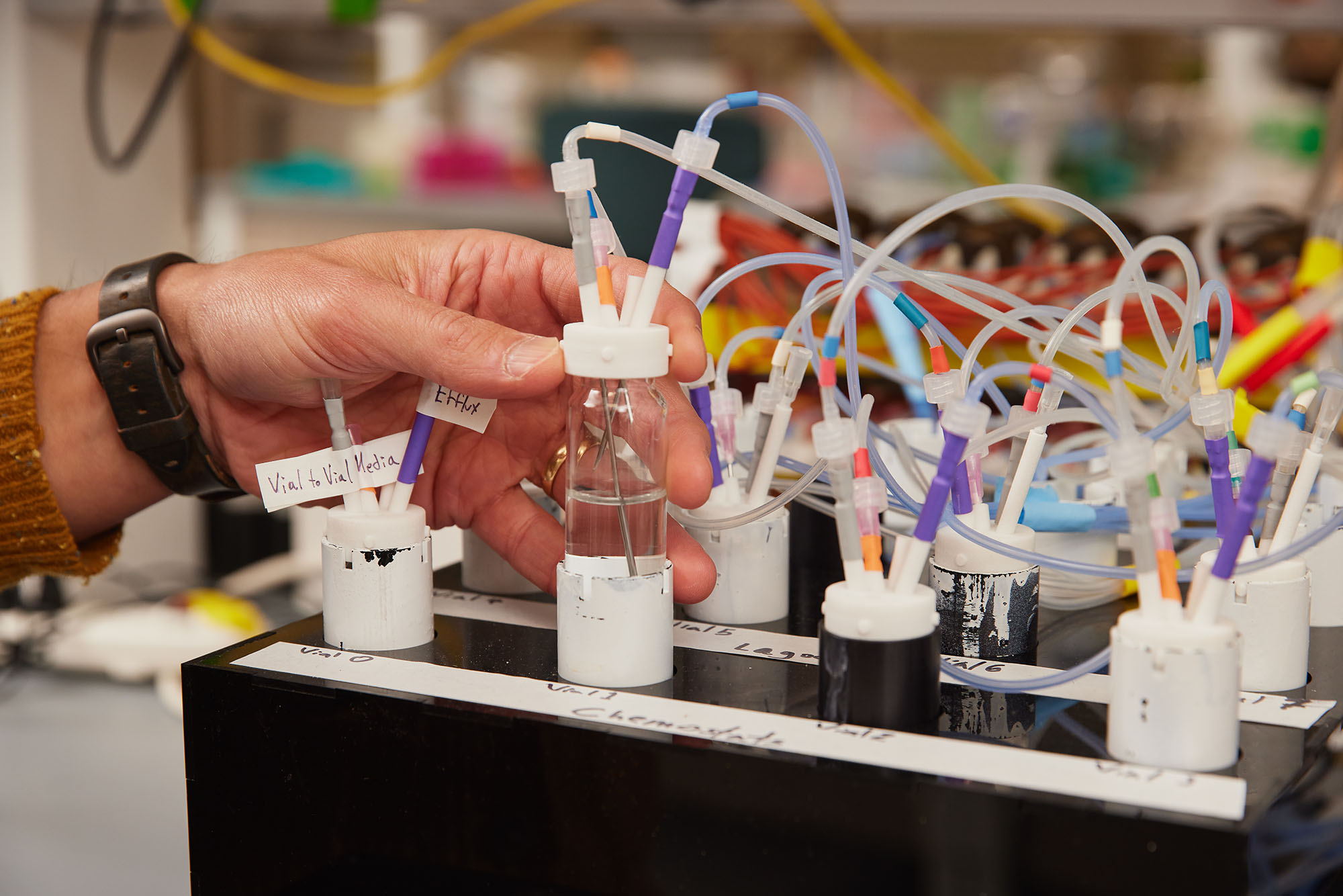 Photo: A hand shows off a test tube with many multicolored wires attached to it in a lab.
