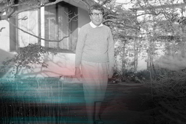 Black and white photo of Writer Nobuko Yoshiya, an older Japanese woman wearing a long sleeved seater over a white collared shirt and long skirt. She poses for a photograph in a small rock garden. A small building can be seen behind her.