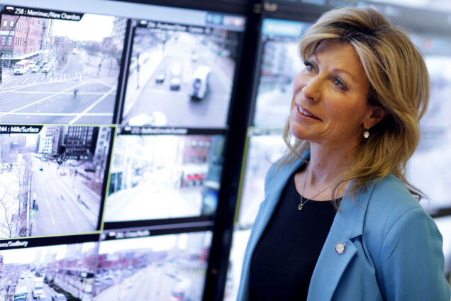 Photo: Gina Fiandaca, a white woman wearing a black shirt and light blue blazer, is shown looking at various screens of video footage of various MBTA stations.