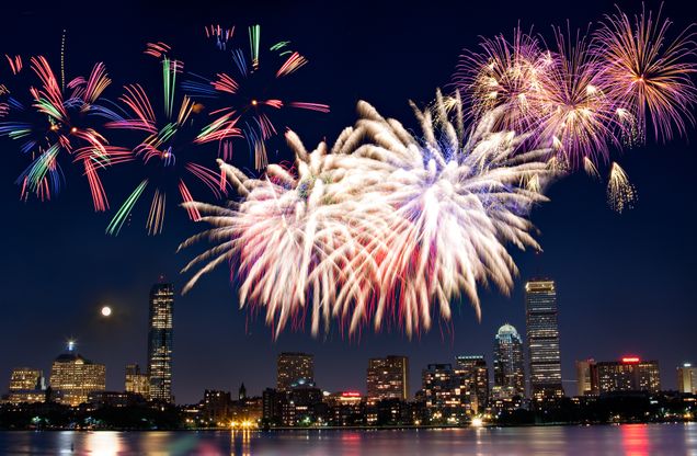 This is a stock image from Canva.com which shows colorful fire works fill the dark skyline in Boston. The photo is taken so you can see the River, as the the multi-colored fireworks reflects off the water. You can see the different sized buildings with their lights on in the background.