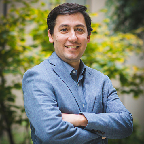 Photo: Hadi T. Nia poses for a photo with arms crossed. He wears an ocean blue blazer and smiles for the camera in front of a blurry landscape of plants.