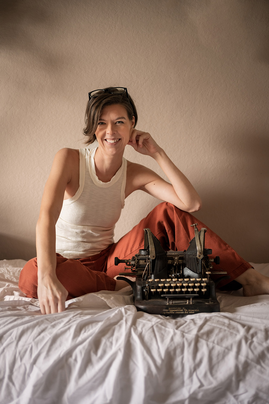 Photo: Jillian Abby, a white woman with short, brown hair and wearing a white tank top and orange-red casual pants, smiles and poses with one elbow resting on her knee. She sits on a white bed in front of a black, old school typewriter.