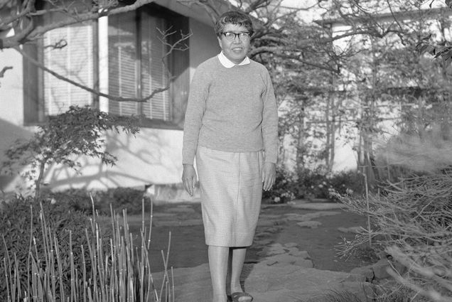 Black and white photo of Writer Nobuko Yoshiya, an older Japanese woman wearing a long sleeved seater over a white collared shirt and long skirt. She poses for a photograph in a small rock garden. A small building can be seen behind her.