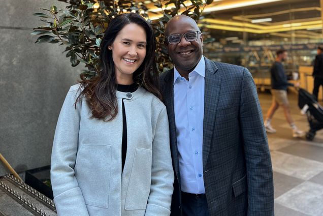 Photo: Arianne Kidder (left) stands and poses next to Jason Robart. A white woman with dark brown hair and wearing a black blouse and light grey jacket smiles and poses next to a bald, Black man wearing glasses, a light blue button-up collared shirt, and dark grey blazer.