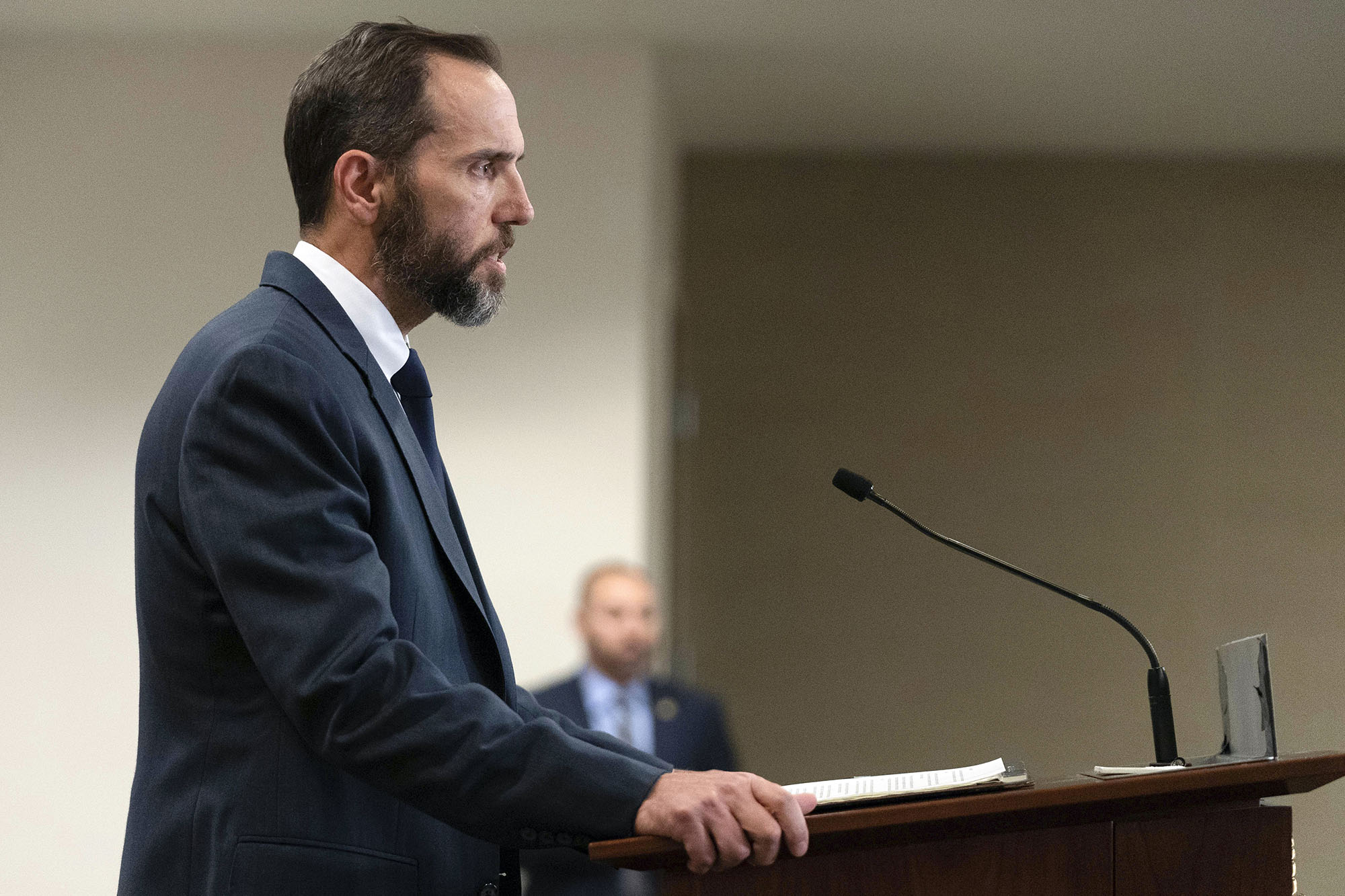 Photo: Special counsel Jack Smith, a white man with dark hair and beard and wearing a black suit, is shown speaking to reporters at a wooden podium with small microphone.