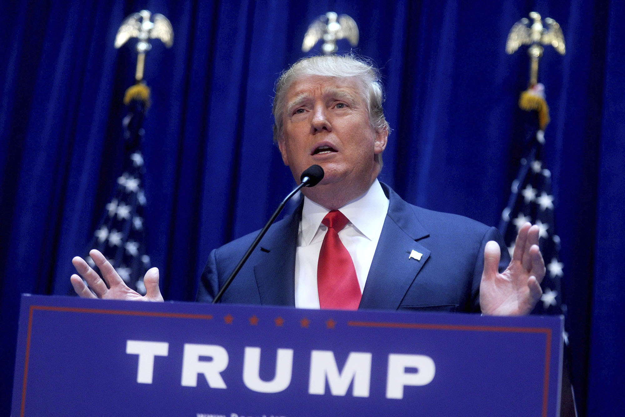 Photo: Donald Trump, an older white man with thinning silver hair and an orange tan and wearing a navy suit and red tie, is shown mid-talk as he holds his hands up palms forward at a blue podium and microphone. Text on podium reads "Trump". Three American flags are positioned behind him.