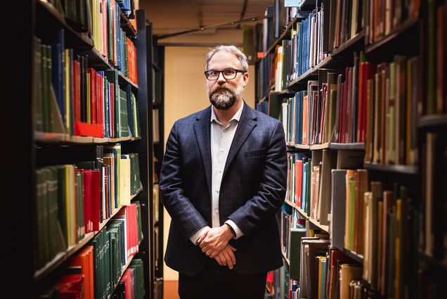 Photo: Mark Newton, a white man with dark and light gray hair, beard, and mustache and wearing glasses, a light blue collared shirt, and navy blue blazer and pants, poses with hands clasped in front of him between two large large library shelves filled with books.